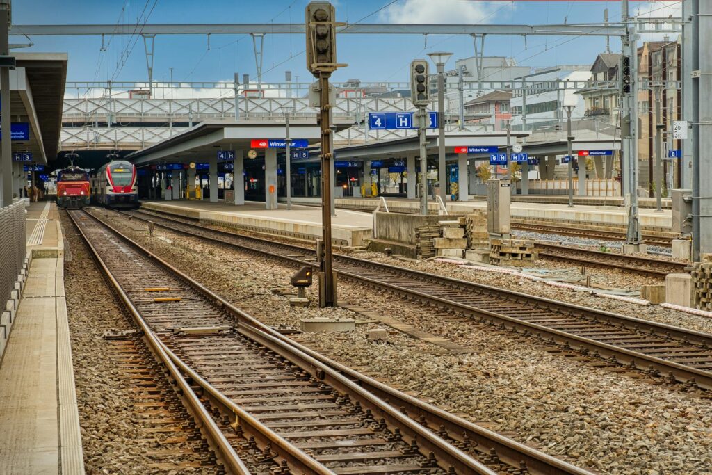 A wide-angle view of Winterthur train station in Switzerland, showing multiple railway tracks converging toward platforms. Two modern trains are stationed on the left side, and overhead signage clearly marks platforms 3 to 7. The station infrastructure includes overhead power lines, signal lights, and a pedestrian bridge above the platforms. The surrounding area has a mix of modern and traditional European-style buildings under a blue sky with scattered clouds.